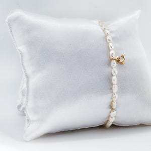 May include: A delicate pearl bracelet with a small gold charm featuring a clear gemstone. The bracelet is displayed on a white satin pillow.