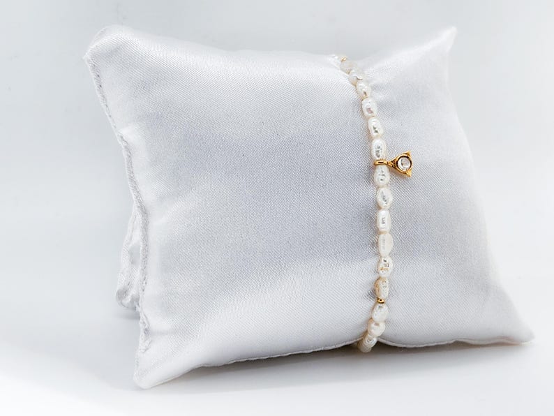 May include: A delicate pearl bracelet with a small gold charm featuring a clear gemstone. The bracelet is displayed on a white satin pillow.