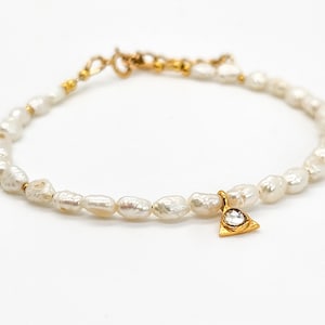 May include: A delicate pearl bracelet with a gold triangle charm featuring a clear gemstone. The bracelet is made of small, irregular pearls and is perfect for adding a touch of elegance to any outfit.