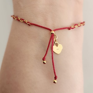 May include: Red and gold bracelet with a heart charm. The bracelet features a red cord with gold chain accents and a small gold heart pendant. The heart charm has the number "375" engraved on it. The bracelet is adjustable.