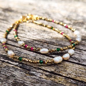 May include: Two gold-colored beaded bracelets with white, oval-shaped pearls and small red and green beads. The bracelets are on a weathered wooden surface, creating a natural and elegant aesthetic. Jewelry.