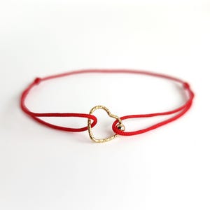 Peut inclure: Un bracelet en cordon rouge avec un pendentif en forme de cœur en or. Le pendentif en forme de cœur a une surface texturée.