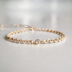 Puede incluir: Pulsera dorada con una gema transparente en el centro. La pulsera presenta pequeños eslabones circulares y un cierre de nudo corredizo. El diseño es sencillo y elegante, adecuado para el uso diario.