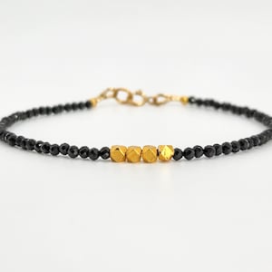 May include: A delicate bracelet featuring small, black beads interspersed with five gold-coloured, cube-shaped beads. The bracelet has a gold-coloured clasp and is displayed against a white background. This piece of jewellery is suitable for everyday wear.
