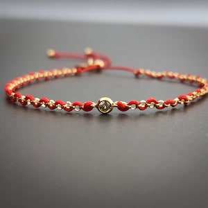 May include: Red and gold bracelet with a central clear gemstone. The bracelet features a red cord with gold-colored chain links and a sliding knot closure. A delicate piece of jewelry.
