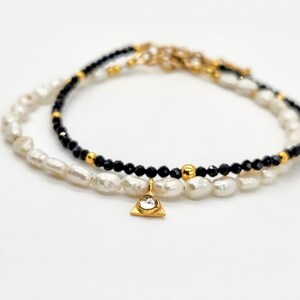 May include: A delicate layered bracelet featuring black beads and white pearls with a gold triangle charm accented with a clear rhinestone.
