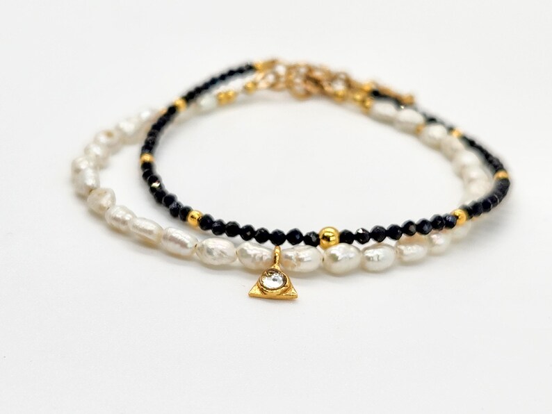 May include: A delicate layered bracelet featuring black beads and white pearls with a gold triangle charm accented with a clear rhinestone.