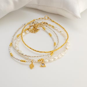 May include: Set of four delicate gold and silver beaded bracelets with pearl accents and two small charms. The charms are a diamond shape and a triangle with a clear stone in the center.