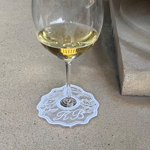 May include: A clear glass wine glass filled with a pale yellow liquid, featuring a white lace coaster with a scalloped edge and the embroidered monogram letters KB placed around the base of the stem on a flat, light-colored stone surface.