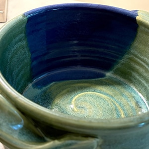 May include: A handmade ceramic bowl with a glossy finish. The bowl features a blend of colors, including shades of blue, green, and a hint of yellow. The interior has a swirling pattern, and the exterior has two handles.