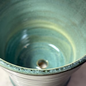 May include: A handcrafted ceramic planter with a turquoise and green glaze. The interior features a drainage hole at the bottom. The rim has a textured, speckled finish. The planter's design is a wide, open bowl shape.