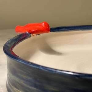 May include: A ceramic bowl with a smooth off-white interior and a dark blue exterior featuring visible brushstroke textures. A small, bright red sculpted bird figure is attached to the top edge of the rim, extending outward from the bowl.