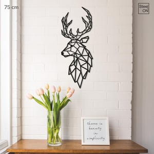May include: Black geometric deer head wall decor made of metal. The deer head is 75 cm tall and is mounted on a white brick wall. There is a vase of pink tulips and a framed quote that reads "There is beauty in simplicity" on a wooden table in front of the wall.