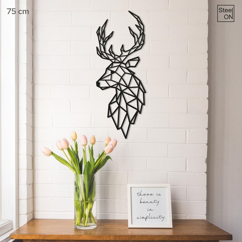 May include: Black geometric deer head wall decor made of metal. The deer head is 75 cm tall and is mounted on a white brick wall. There is a vase of pink tulips and a framed quote that reads "There is beauty in simplicity" on a wooden table in front of the wall.