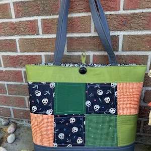 May include: A green and black patchwork tote bag with a skull print. The bag has a gray handle and a green button closure.