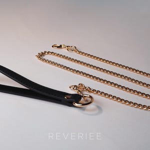 May include: A black leather handle with a gold-tone ring and a gold-colored chain leash. The chain is made of small, interlocking links. The word "REVERIEE" is printed in white at the bottom of the image.