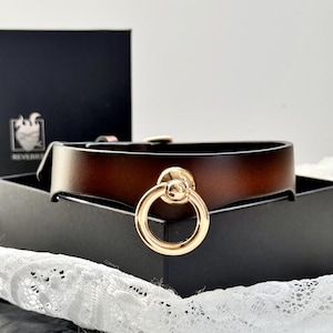 May include: A brown leather choker with a gold-toned ring and a small ball accent. The choker is on a black box with a white lace fabric in the background.