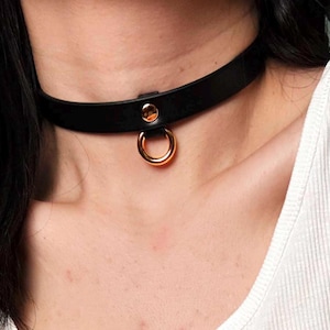 May include: A black choker necklace with a gold-colored ring and rivet detail. The choker is made of a smooth, dark material and is worn around the neck. The gold ring hangs from the center of the choker.