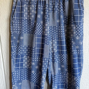 May include: Blue patchwork shorts with an elastic waistband. The shorts feature a variety of white geometric patterns, including squares, circles, and stripes. The shorts are hanging on a gold-colored hanger.