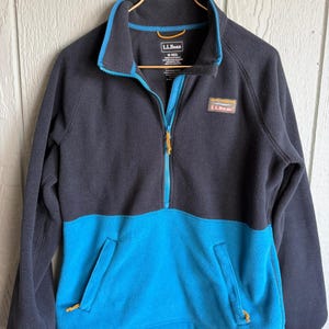 May include: A navy blue and turquoise fleece jacket with a half-zip closure. The jacket features a turquoise collar and zipper trim, two front pockets, and the "L.L.Bean" logo patch. The jacket is hanging on a gold-colored hanger.