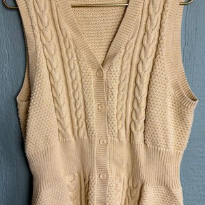 Könnte beinhalten: Eine cremefarbene Strickweste mit Zopfmuster, Knopfleiste und zwei Taschen. Die Weste ist aus einem weichen, bequemen Material gefertigt und hat eine lockere Passform.