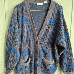 May include: A vintage button-down cardigan in shades of blue, gray, and black, featuring a textured knit design. The sweater has a V-neck, two front pockets, and a blue paisley pattern. The label reads "Wanderlust".
