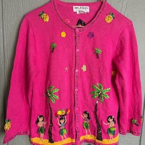Puede incluir: Un cárdigan rosa brillante con diseños tropicales bordados. El suéter presenta palmeras, piñas y bailarinas de hula. El texto "Jack B. Quick" está en una etiqueta cerca del escote. Tiene una parte delantera abotonada y mangas largas.