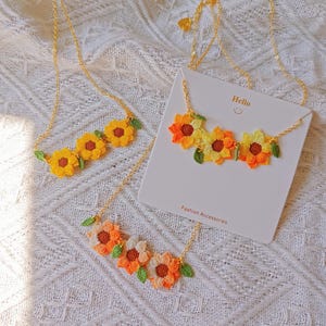 Può includere: Collane di girasoli all'uncinetto con catene dorate. Le collane presentano ciondoli a forma di fiori gialli, arancioni e bianchi con foglie verdi. Una collana è esposta su una carta bianca con la scritta "Hello" e "Fashion Accessories".