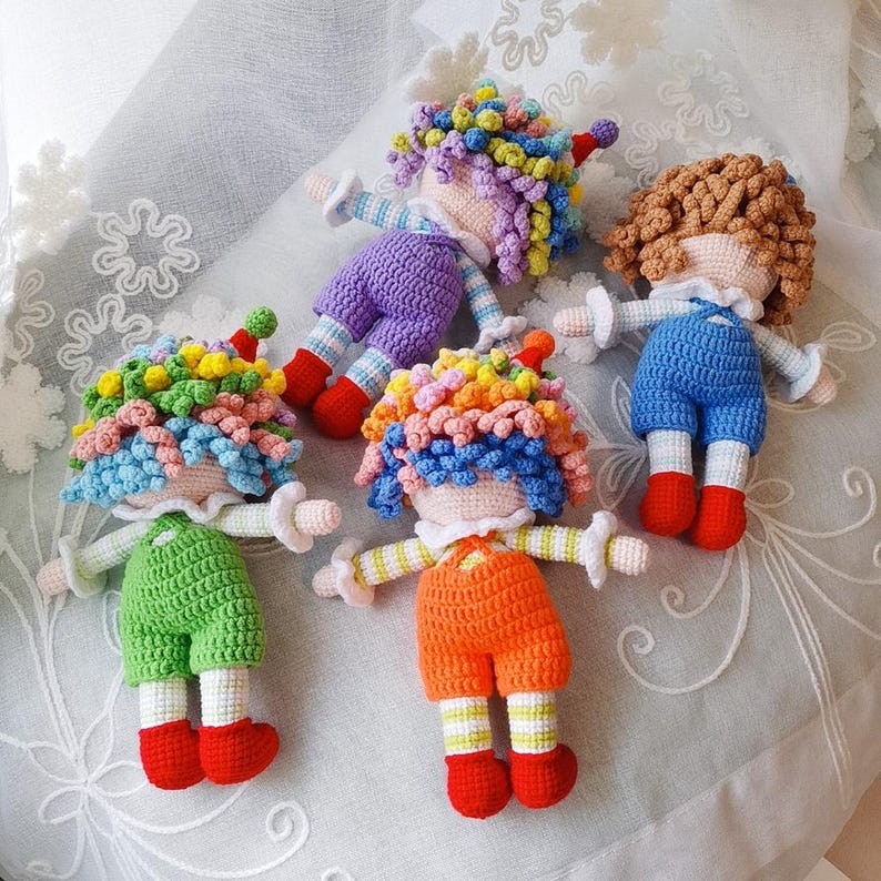 May include: Five handmade crochet dolls with colorful yarn hair, each wearing different colored outfits. The dolls have red shoes and white accents. The outfits are green, orange, blue, and purple.