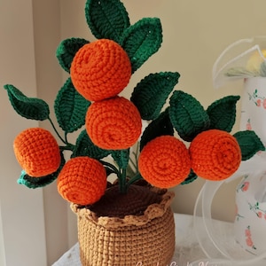 May include: A handmade crocheted orange tree featuring vibrant orange fruit and green leaves, presented in a brown crocheted pot with a decorative, ruffled edge. The text "CrochetCrafty Nest" is visible on the pot.