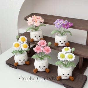Peut inclure: Cinq pots de fleurs crochetés avec des visages souriants et des pieds bruns. Chaque pot contient des fleurs crochetées de couleurs différentes, notamment roses, violettes et blanches avec des centres jaunes. Les pots sont disposés sur une étagère en bois.