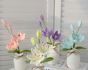 May include: A collection of handmade crochet lily flowers in small white vases. The lilies are in various colors, including pink, white, purple, and blue. Each vase is tied with a brown string. The text "ATABLE IN PARIS" is visible on a book.