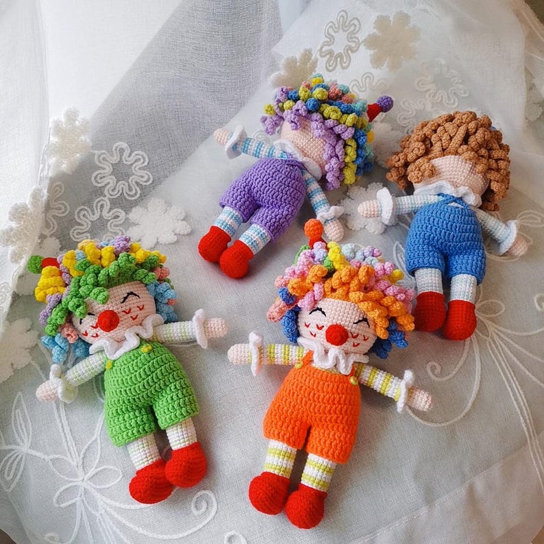 May include: Four handmade crochet clown dolls in various colors. Each doll has a smiling face, a red nose, and colorful yarn hair. The clowns wear different colored outfits, including orange, green, purple, and blue, with red shoes and white accents.