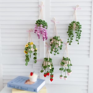 May include: Six crocheted hanging planters with different flowers and greenery. The planters are made of beige yarn and are hanging from white string. The flowers are pink, yellow, and white. The greenery is green.