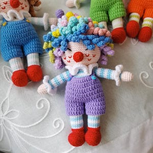 May include: Crocheted clown doll with colorful curly hair, a red nose, and a white ruffled collar. The doll wears a purple jumpsuit, striped sleeves, and red shoes. Other similar dolls are visible in the background.