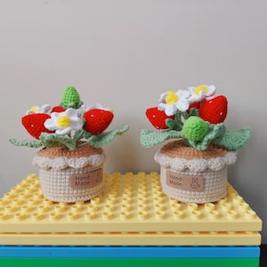 May include: Two crocheted strawberry plants in pots. Each plant has red strawberries, white flowers, and green leaves. The pots are beige and have a brown label that says "Hand Made".
