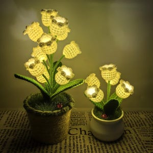 May include: Two decorative flower arrangements with glowing yellow crochet flowers. One is in a woven pot, the other in a white ceramic pot. Both have green leaves and a small ladybug.