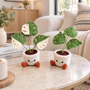 May include: Two crocheted monstera plants in cream-coloured pots with smiling faces and small orange feet. Each plant features green and cream variegated leaves with cutouts, supported by thin green stems, and dark brown soil texture on top.
