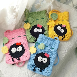 May include: Five colourful, handmade crochet bags. Each bag is a different colour: pink, green, yellow, and blue. They feature a black character with white eyes, a red bow, and decorative flowers. The bags have a loop handle and are displayed on a white surface.