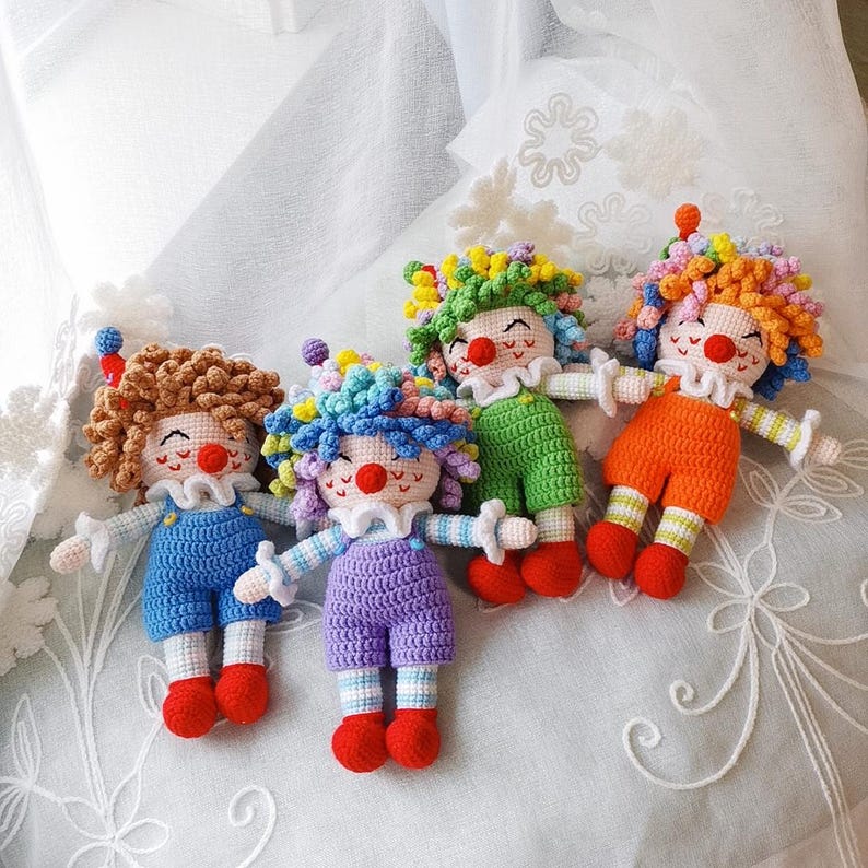 May include: Four handmade crochet clown dolls. Each doll has a colorful, curly wig, a red nose, and a white ruffled collar. The clowns wear blue, purple, green, and orange outfits with red shoes. The dolls have outstretched arms and are smiling.