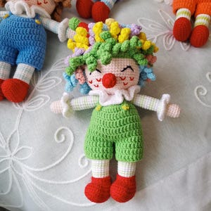 May include: Crocheted clown doll with a red nose, colorful curly hair, and a green jumpsuit. The doll has white ruffled collar and cuffs, and red shoes. The doll is handmade and appears to be part of a collection.