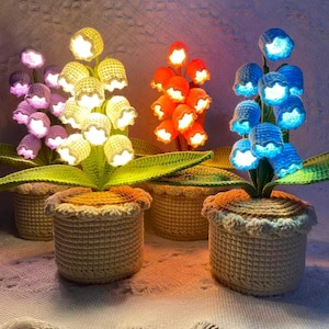 Może przedstawiać: Szydełkowe lampy w kształcie kwiatów w kolorach fioletowym, żółtym, pomarańczowym i niebieskim. Każdy kwiat jest podświetlony, a rośliny znajdują się w pasujących szydełkowych koszach. Lampy są ustawione na teksturowanej powierzchni.