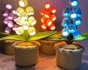 May include: Crocheted flower lamps in various colors, including purple, yellow, orange, and blue. Each flower is illuminated, and the plants are potted in matching crocheted baskets. The lamps are arranged on a textured surface.