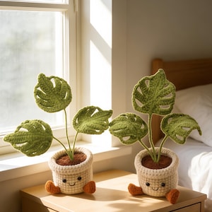 May include: Two crocheted plant decorations with green leaves and stems in cream-coloured pots. The pots have smiling faces and orange feet. The plants sit on a wooden bedside table near a window. Text reads "Crochet Crafty Nest".