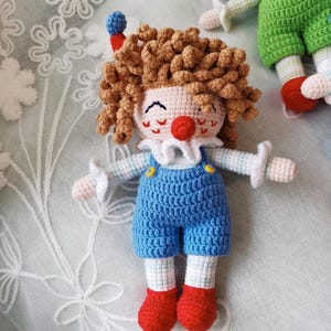 May include: A handmade crochet clown doll with a curly brown hair, a red nose, and a white ruffled collar. The doll wears blue overalls with yellow buttons and red shoes. The doll is a whimsical, handcrafted item.
