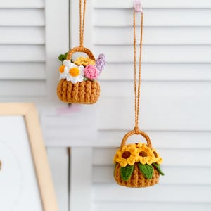 May include: Two crocheted flower baskets hanging from a white slatted wall. The top basket has a mix of white, yellow, pink, and purple flowers. The bottom basket has yellow sunflowers.