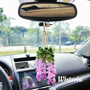 May include: A crocheted wisteria flower hanging from a rearview mirror in a car. The flower is pink and purple with green leaves. The text "Wisteria" is visible in the image.