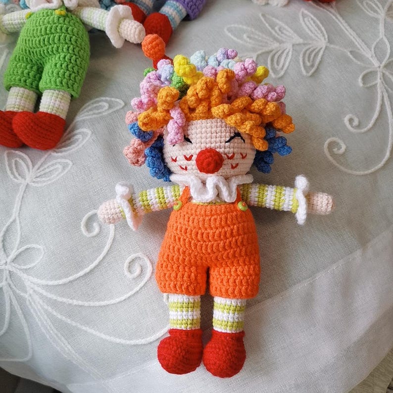 May include: A handmade crochet clown doll with orange shorts, red shoes, and a white ruffled collar. The doll has colorful curly hair, a red nose, and is smiling. The arms are outstretched, and the doll is made with a textured stitch.