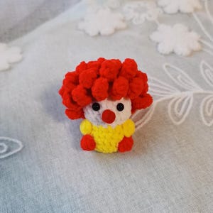 May include: A handmade crochet clown doll with a red curly wig, white face, black eyes, and a red nose. The clown wears a yellow shirt with red accents. The doll is a whimsical, handcrafted item.