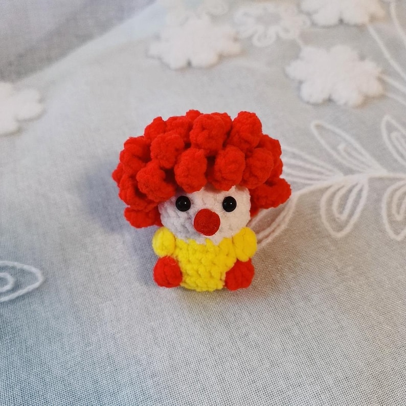 May include: A handmade crochet clown doll with a red curly wig, white face, black eyes, and a red nose. The clown wears a yellow shirt with red accents. The doll is a whimsical, handcrafted item.